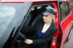 ritratto di bella bionda modello di moda donna sexy in berretto e in tutto nero con trucco luminoso sedersi e guidare la city car rossa. foto