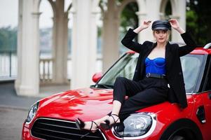 ritratto di bella bionda modella donna moda sexy in berretto e in tutto nero con trucco luminoso sedersi sul cofano della city car rossa. foto