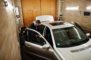 uomo in uniforme e respiratore, lavoratore del centro di autolavaggio, pulizia della spazzola per la pulizia degli interni dell'auto. concetto di dettagli dell'auto. foto