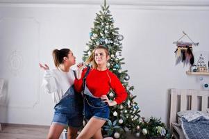 due divertenti amiche di belle ragazze indossano in tute, pantaloncini di jeans e ghette contro l'albero di Capodanno con decorazioni natalizie. foto