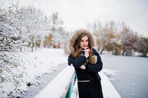 bella ragazza bruna in abiti caldi invernali. modello su giacca invernale contro il lago ghiacciato al parco. foto