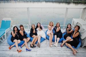 un gruppo di 8 ragazze indossano abiti neri e 2 spose alla festa di addio al nubilato seduti sui cuscini al molo sul lato della spiaggia. foto