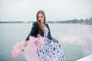 la ragazza alla moda in giacca di pelle tiene il bagliore di fumo rosa al giorno d'inverno contro il lago ghiacciato. foto