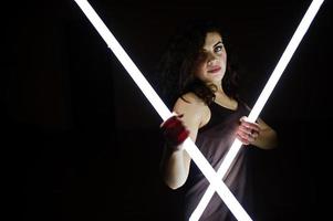 bruna riccia modello plus size con due lunghe lampade a led in camera nera. foto