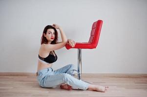 ragazza dai capelli rossi con un grande busto su reggiseno nero e jeans seduta vicino a una sedia rossa contro il muro bianco in una stanza vuota. foto