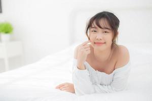 ritratto bella giovane donna asiatica sorriso mentre sveglia sano e benessere al mattino in camera da letto, bellezza asia ragazza sdraiata cura della pelle e trucco cosmetico con felice, stile di vita e relax concetto. foto