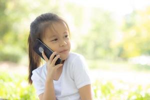 bellissimo ritratto bambino asiatico felice e parlando telefono cellulare intelligente nel parco naturale in estate, cellulare asiatico bambino, bambina casual relax e tempo libero, concetto di comunicazione. foto