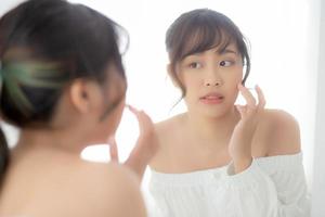 ritratto di bellezza giovane donna asiatica sorridente sguardo allo specchio di controllo della cura della pelle caucasica con benessere in camera da letto, bella ragazza felice faccia commovente nella riflessione per la salute, concetto di stile di vita. foto