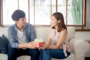 la giovane coppia asiatica festeggia il compleanno insieme, l'uomo asiatico regala una confezione regalo alla donna per una sorpresa in soggiorno, l'anniversario felice ed eccitato femminile, il concetto di San Valentino per le vacanze. foto