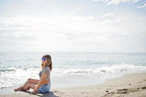 bellissima modella che si rilassa su una spiaggia di mare, indossa jeans corti, camicia leopardata e occhiali da sole. foto