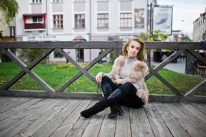 ragazza bionda al cappotto di pelliccia che si siede sul pavimento di legno all'aperto. foto