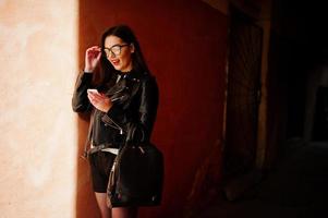 elegante ragazza bruna indossare su giacca di pelle e pantaloncini con zaino e occhiali contro la parete arancione alle ombre guardando sul cellulare. foto