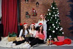 quattro ragazze carine amiche indossano maglioni caldi, pantaloni neri e cappelli di Babbo Natale contro l'albero di Capodanno con decorazioni natalizie in studio. foto
