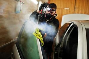 uomo in uniforme e respiratore, lavoratore del centro di autolavaggio, pulizia degli interni dell'auto con pulitore a vapore caldo. concetto di dettagli dell'auto. foto