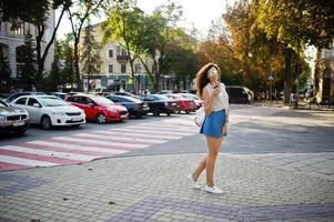 la ragazza alla moda riccia indossa la gonna, la camicetta e gli occhiali dei jeans blu poste in una strada della città con una tazza di caffè. foto