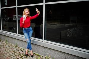 ritratto di una bella donna in camicetta rossa e jeans casual che si fa selfie sul cellulare e tiene una tazza di caffè fuori dall'enorme centro commerciale. foto