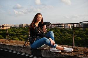 ritratto di una bella giovane donna in giacca di pelle nera, jeans e scarpe da ginnastica seduta su un tetto con una vista incredibile sullo sfondo. foto