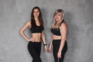 due amiche con diversi tipi di corpo e in abbigliamento sportivo in piedi in studio con sfondo grigio foto