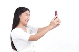 ritratto di bella donna asiatica che ha i capelli lunghi neri in camicia bianca, tiene lo smartphone in mano e sorride. lei scatta una foto di selfie su sfondo bianco.