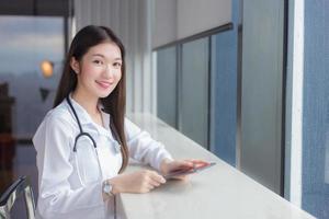 la dottoressa professionista asiatica vestita di camice bianco si siede su una sedia in una biblioteca ospedaliera per cercare alcune informazioni su un tablet per il trattamento nel concetto di assistenza sanitaria. foto