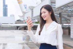 la giovane donna d'affari asiatica in abito bianco sorride e tiene uno smartphone in mano per la videochiamata in una felice giornata lavorativa nella città all'aperto come sfondo. foto
