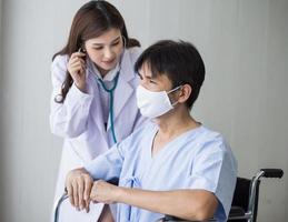 la dottoressa asiatica usa uno stetoscopio per controllare il ritmo polmonare di un paziente maschio che indossa una maschera facciale mentre si siede su una sedia a rotelle in ospedale. foto