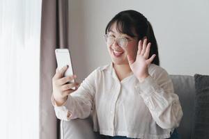 giovane donna asiatica che utilizza lo smartphone per la videoconferenza online agitando la mano facendo un gesto di saluto sul divano in soggiorno. foto