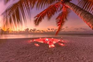 cena romantica in spiaggia con tramonto, candele con foglie di palma e tramonto cielo e mare. vista straordinaria, luna di miele o paesaggio per la cena di anniversario. orizzonte serale dell'isola esotica, romanticismo per una coppia foto