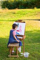 una bambina è seduta sulla panca di legno e dipinta sulla tela posta su un leggio da disegno foto