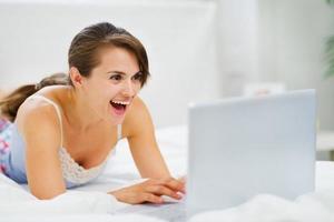 donna che pone sul letto e che osserva sorprendentemente in computer portatile foto