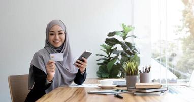bella giovane donna musulmana con l'hijab si siede alla scrivania del suo ufficio, con in mano uno smartphone e una carta di credito. pagamento online, internet banking, concetto di shopping online. immagine ravvicinata foto