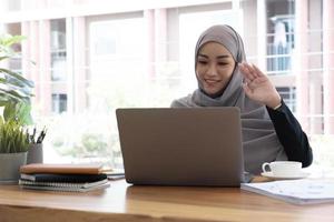 donna d'affari in hijab che ha una chat video sul laptop mentre è seduto al bar. donna seduta al bar e fare videochiamate utilizzando auricolari e computer portatile. foto