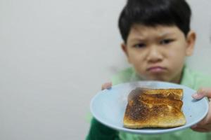 ragazzo asiatico mostra su piatto di pane grigliato bruciato arrosto con faccia di cattivo umore infelice - concetto di delusione della colazione alimentare foto