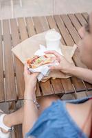 primo piano delle mani della donna che tengono hamburger, donna che mangia fast food al caffè di strada, vista da dietro foto