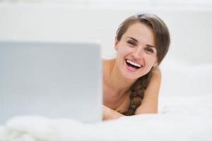 donna sorridente che guarda fuori dal computer portatile mentre giaceva a letto foto