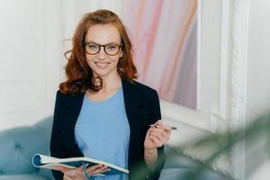 sorridente donna dai capelli rossi scrive appunti nel blocco note, tiene diario e penna, indossa un abito formale, occhiali, posa al chiuso, ascolta consigli, registra nel portafoglio personale, si prepara per la riunione foto