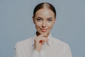 La donna d'affari attraente e sicura di sé tiene il mento e guarda direttamente la fotocamera, indossa un'elegante camicia bianca, ha una pelle sana e un trucco naturale, isolata su sfondo blu. donne, affari, carriera foto