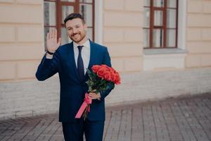 uomo felice vestito formalmente con bouquet di rose e salutando la donna amata all'aperto foto