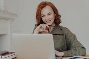 una donna d'affari professionista di successo con i capelli rossi lavora in remoto su un computer portatile, legge articoli sugli affari online, si siede alla scrivania, utilizza tecnologie moderne, prepara un rapporto finanziario foto