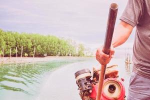 foto in stile vintage di un pescatore sta controllando il motore del timone su una barca nel monte del fiume vicino al mare con la foresta di mangrovie e lo sfondo del cielo blu