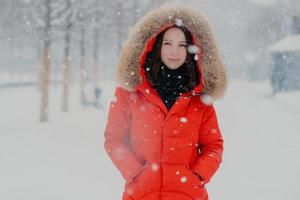 inquadratura alla vita di una donna sorridente attraente con pelle sana, capelli scuri, indossa una giacca rossa, tiene le mani in tasca durante il gelo, si trova su uno sfondo bianco di neve, gode di forti nevicate foto