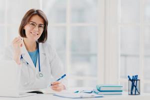 foto di stagista lavora in ospedale, scrive nel certificato medico, prescrive le cure per il paziente, fa la ricetta, vestita di camice bianco con fonendoscopio, lascia appunti di visita medica