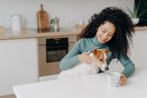 felice donna afro africana con l'acconciatura riccia tratta il cane in cucina, posa al tavolo bianco con una tazza di bevanda, goditi l'atmosfera domestica, fai colazione insieme. persone, animali, concetto di casa. foto