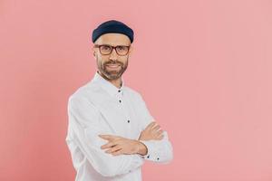 imprenditore maschio positivo con espressione facciale soddisfatta e sicura di sé, tiene le mani incrociate, sta di profilo, guarda direttamente la telecamera, posa in interni su sfondo rosa con spazio libero foto