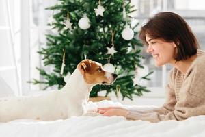 inquadratura orizzontale di bella donna allegra gode di stare insieme con il cane, tiene le sue zampe, sdraiarsi sul letto bianco, guardarsi negli occhi, sentire amore e amicizia, albero di natale decorato sullo sfondo foto