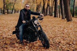 concetto di viaggio, velocità, libertà. il motociclista maschio alla moda posa su una moto nera, indossa occhiali da sole protettivi, giacca e scarpe nere, cavalca nel bellissimo parco giallo durante il periodo autunnale. foto