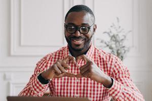 uomo africano felice che gesturing la forma del simbolo del cuore con le mani durante la videochiamata con la famiglia foto