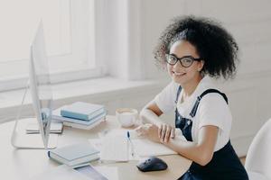 una libera professionista afroamericana positiva posa sul posto di lavoro con documenti e libri di testo, lavora in remoto al computer, fa una pausa caffè, lavora nel suo armadietto, indossa abiti casual. persone, lavoro foto