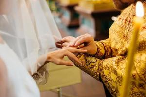 vista del sacerdote che indossa l'anello sulla mano della sposa, posa nella chiesa cristiana durante la cerimonia di matrimonio, candela accesa in primo piano foto