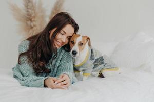 foto di bella donna bruna in maglione lavorato a maglia giace insieme con il cane su un letto morbido, ama passare il tempo con l'animale preferito, si prende cura degli animali, resta a casa durante la quarantena del coronavirus